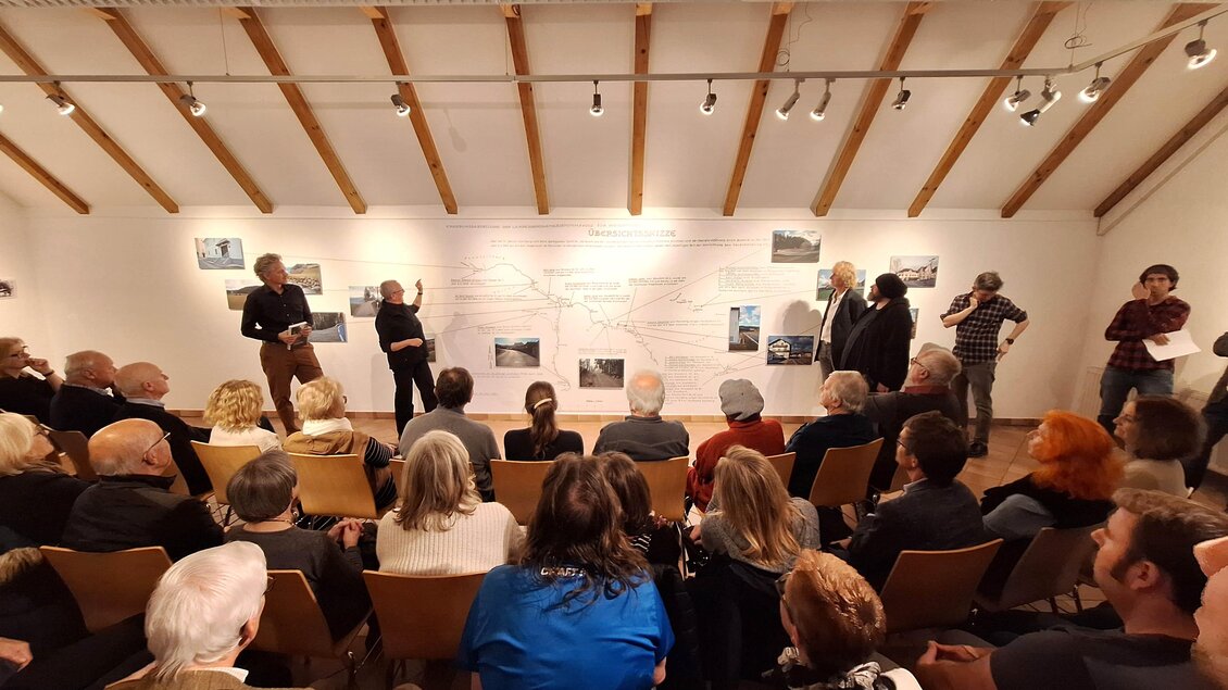 Eine Gruppe von Menschen bei einer Präsentation in einem heller Raum. Im Hintergrund sind Bilder an der Wand zu sehen.