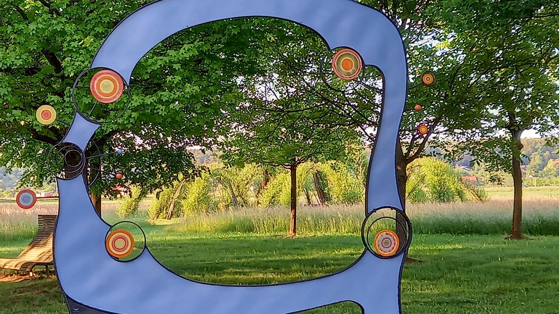 Eine moderne Skulptur in Form eines Rahmens steht in einer grünen Parklandschaft. Bunte Kreise sind in die Skulptur integriert und verleihen ihr ein kreatives Aussehen. | © Thermen- & Vulkanland