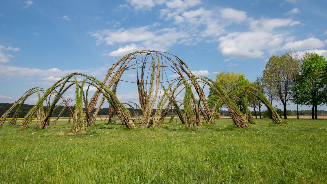Eine moderne Skulptur aus Weiden, die in einem grünen Feld steht. Im Hintergrund sind Bäume und ein blauer Himmel zu sehen. | © Kurkommission Bad Blumau