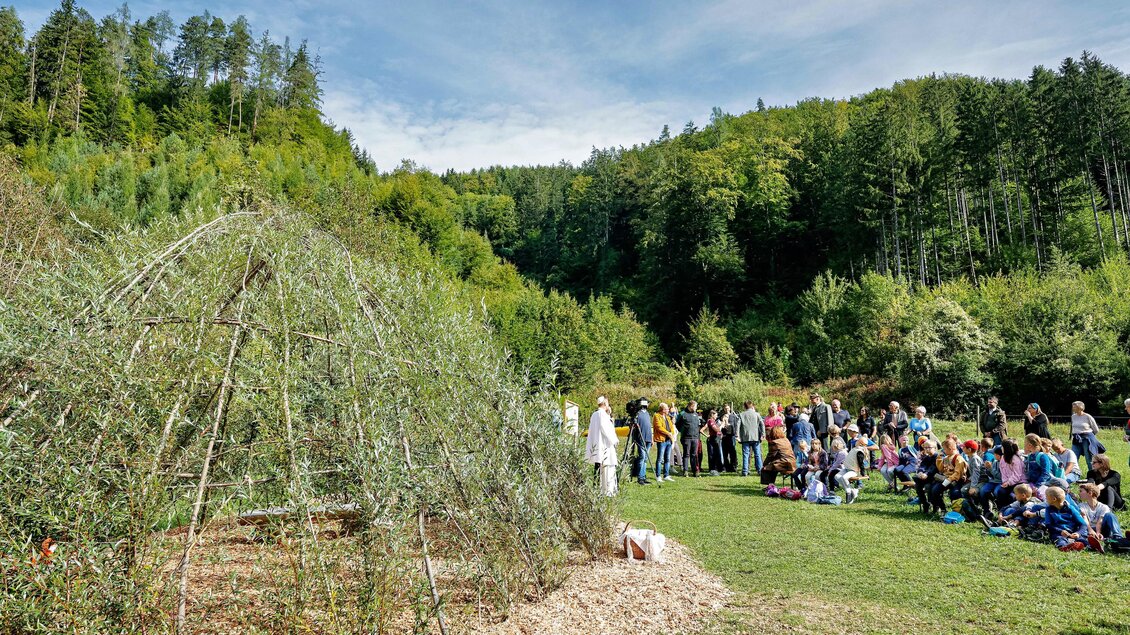 Eine Gruppe von Menschen versammelt sich im Freien, um eine Veranstaltung zu sehen. Im Vordergrund befindet sich eine grüne Struktur aus Zweigen, umgeben von Bäumen und Wiese. | © Gerd Neuhold