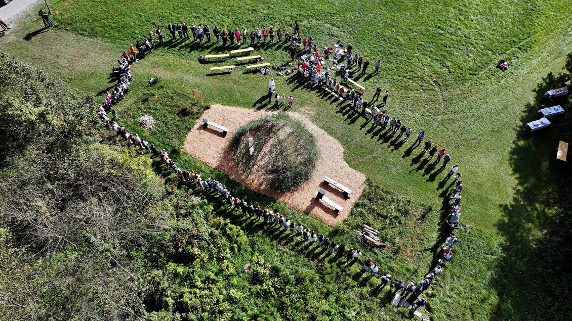 Eine große Gruppe von Menschen steht in einer Linie auf einer Wiese. In der Mitte steht eine runde Struktur, umgeben von Holz und Gras. | © Gerd Neuhold