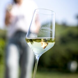 Ein Weinglas mit weißem Wein steht im Vordergrund. Im Hintergrund ist eine Person auf einem Weinberg zu sehen. | © TVB Südsteiermark