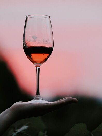 A glass of rosé wine rests on a hand. In the background, a beautiful sunset illuminates the sky. | © TVB Südsteiermark - Lupi Spuma