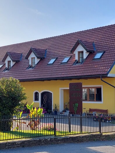 A large, yellow house with red roof tiles and windows in the attic. The garden is green and well-maintained, with a cozy bench and a fence around the property. | © CZ