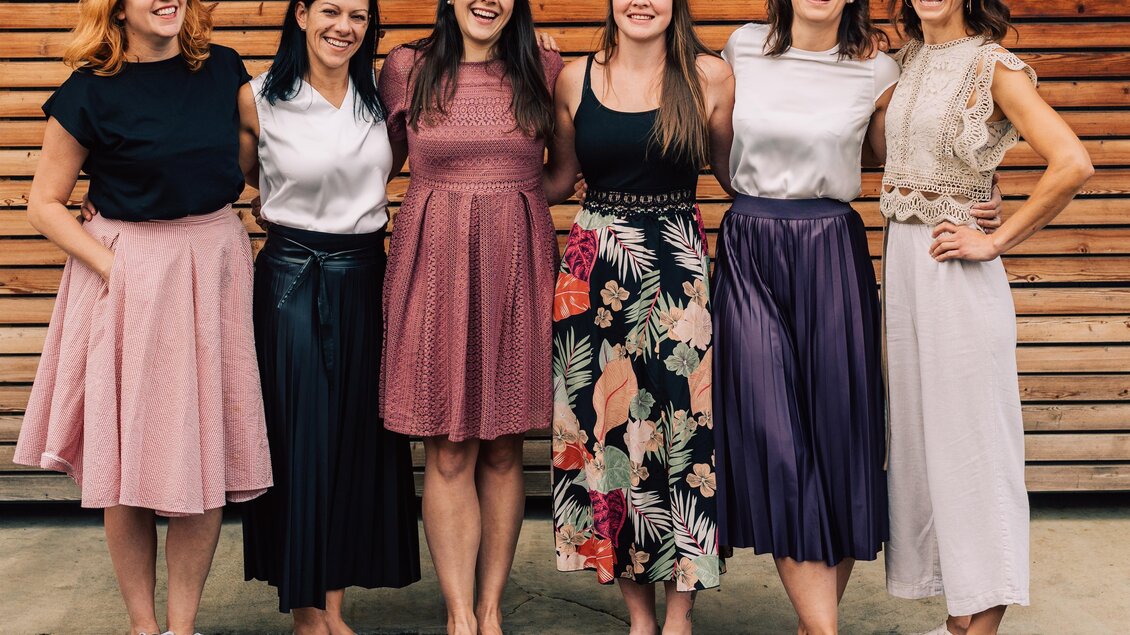 Sechs Frauen stehen in einer Reihe vor einer Holzwand. Sie tragen verschiedene Röcke und lächeln freundlich in die Kamera.