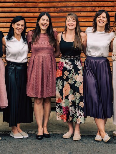 Sechs Frauen stehen in einer Reihe vor einer Holzwand. Sie tragen verschiedene Röcke und lächeln freundlich in die Kamera.