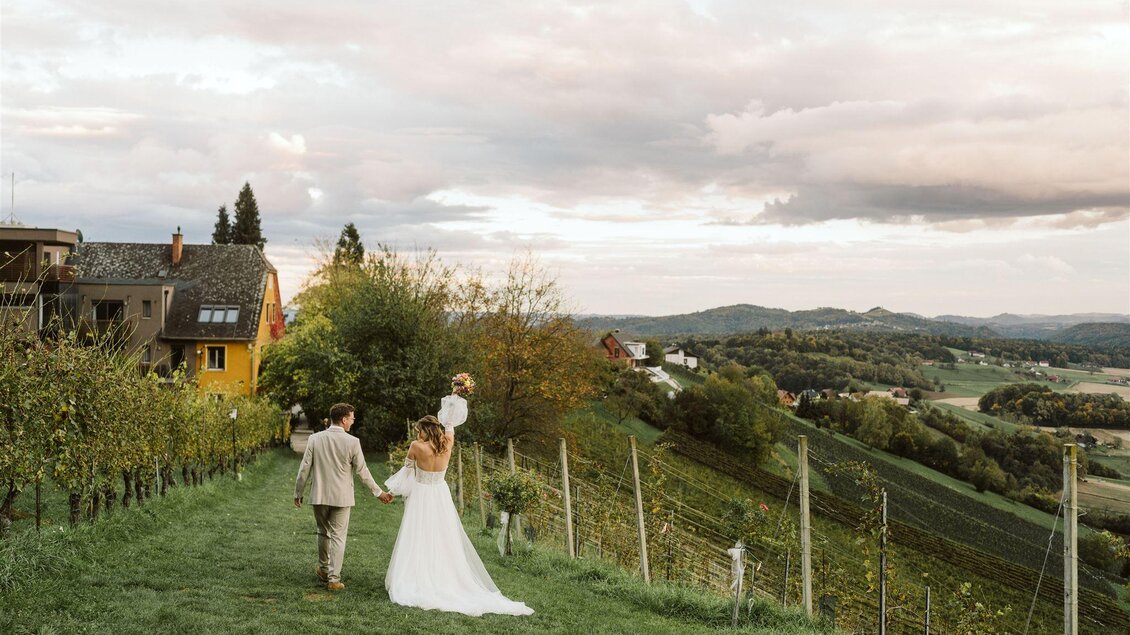 Ein Brautpaar spaziert Hand in Hand durch ein malerisches Weinbaugebiet. Im Hintergrund sind sanfte Hügel und ein bewölkter Himmel zu sehen. | © Weingut Harkamp