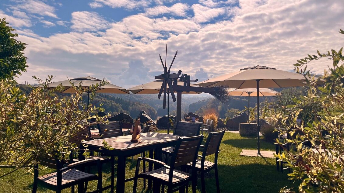 Eine gemütliche Terrasse mit Tisch und Stühlen, umgeben von grünen Pflanzen. Im Hintergrund sieht man Sonnenschirme und eine schöne Aussicht unter einem bewölkten Himmel. | © Michaela Lorber