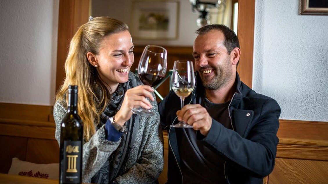Ein Paar stößt mit Weingläsern an und lächelt fröhlich. Im Hintergrund steht eine Flasche Wein auf dem Tisch. | © TV Südsteiermark | Stephan Friesinger