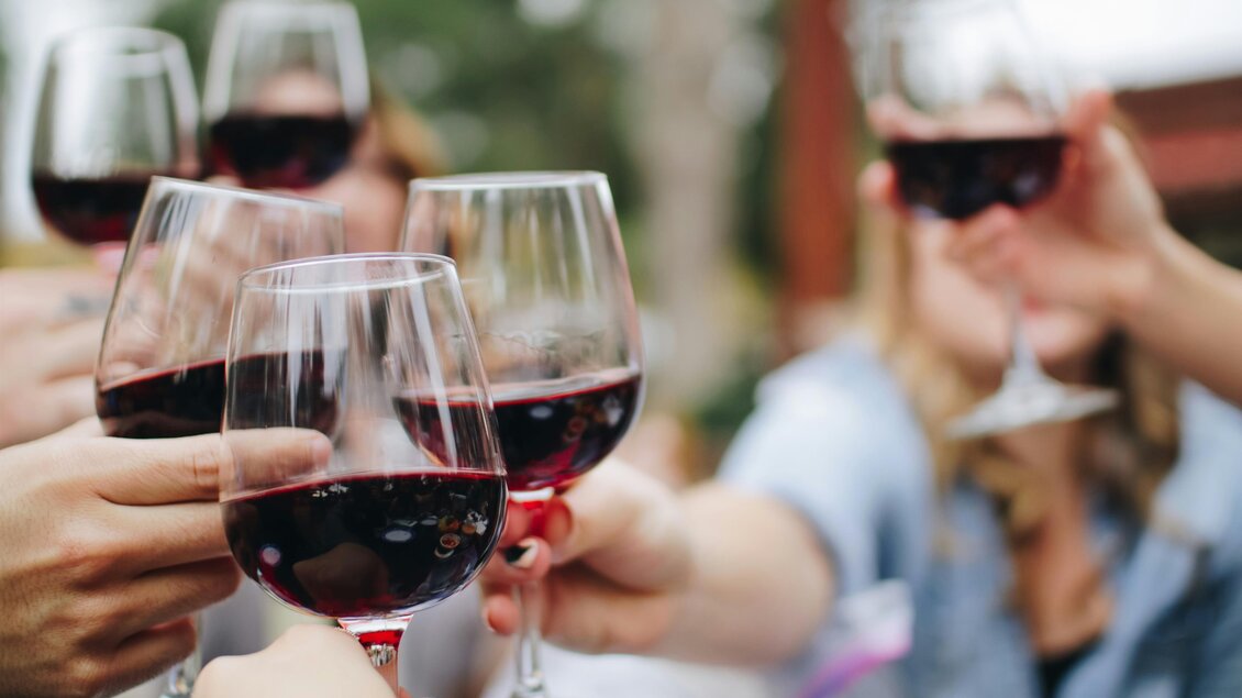 Mehrere Personen stoßen mit Rotwein in Gläsern an. Der Hintergrund zeigt unscharfe Figuren und Pflanzen. | © kelsey-knight-unsplash