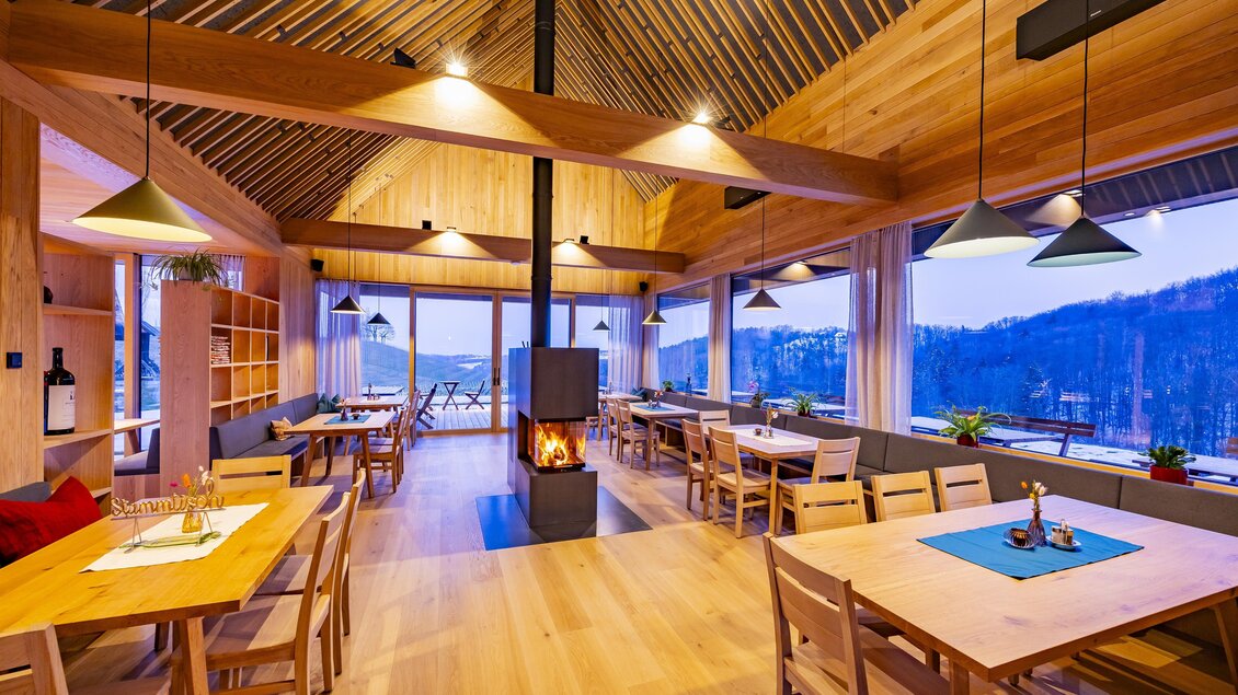 Ein modernes Restaurant mit Holzdekor und großen Fenstern bietet einen Blick auf die Natur. Der zentrale Kamin sorgt für eine gemütliche Atmosphäre. | © BU Dietrich