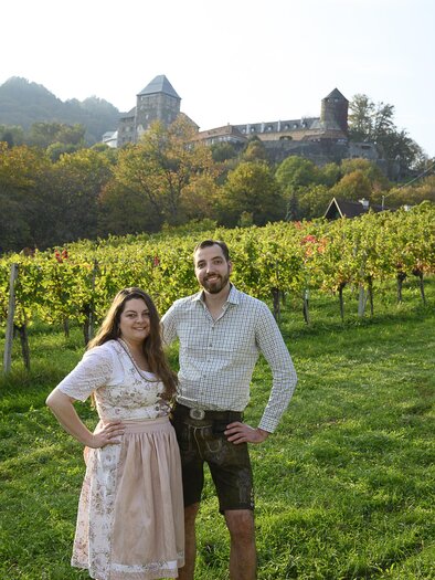 A couple stands in a vineyard with a castle ruin in the background. The woman is wearing a traditional dirndl, and the man is wearing lederhosen.