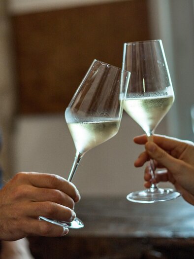 Two glasses of sparkling wine clink together. In the background, blurred, cozy interiors are visible. | © stephan friesinger