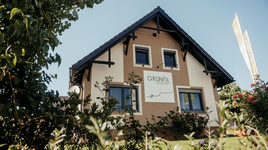 Ein modernes Haus mit einer markanten Fassade und einem Schild, das "GRUNDL" zeigt. Umgeben von grünen Pflanzen und einem klaren Himmel. | © Michaela Lorber