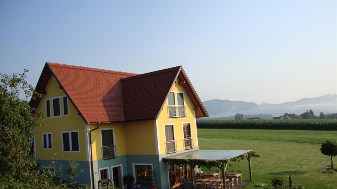 Ein sonniges, gelbes Haus mit rotem Dach steht in einer grünen Landschaft. Im Hintergrund sind sanfte Hügel und ein klarer Himmel zu sehen. | © Winzerhof Grebenz