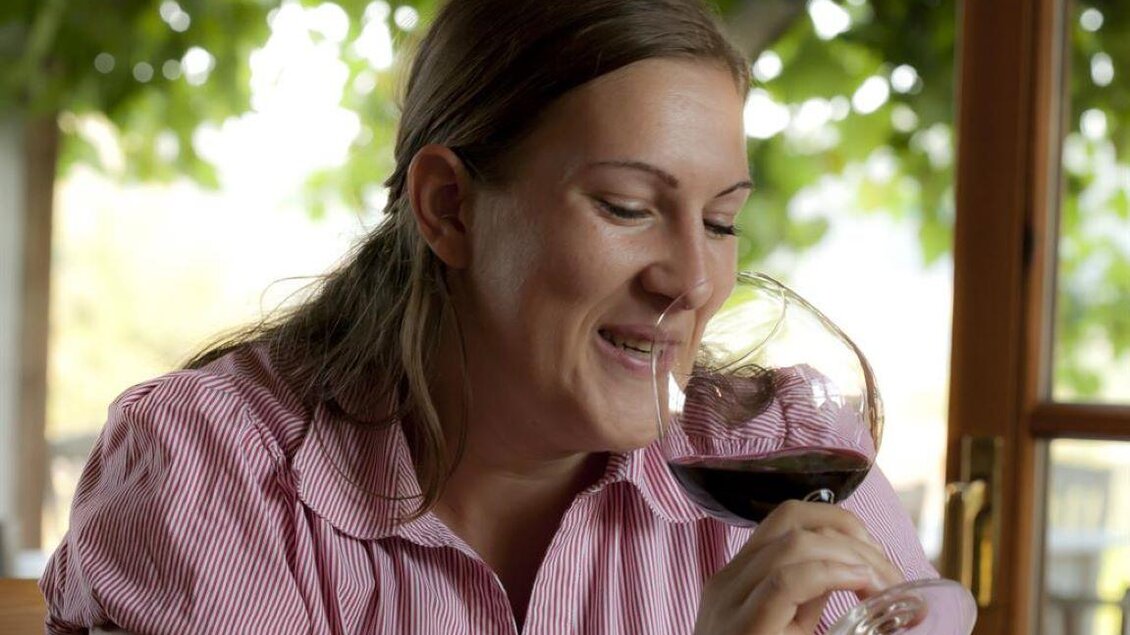 Eine lächelnde Frau genießt ein Glas Rotwein. Im Hintergrund sind Weinreben und ein schöner Ausblick zu sehen. | © Winzerhof Grebenz