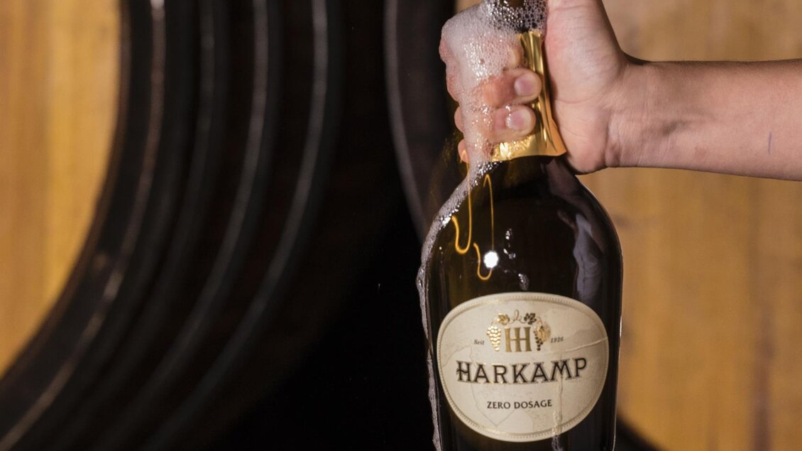 Eine Hand hält eine Flasche Harkamp Zero Dosage, aus der Schaum herausventiliert. Im Hintergrund sind große Holzfässer sichtbar. | © Weingut Hannes und Petra Harkamp