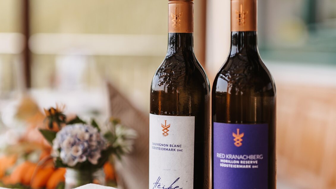 Zwei Flaschen Wein stehen auf einem Holztisch. Im Hintergrund sind Blumen und eine natürliche Landschaft zu sehen. | © Michaela Lorber