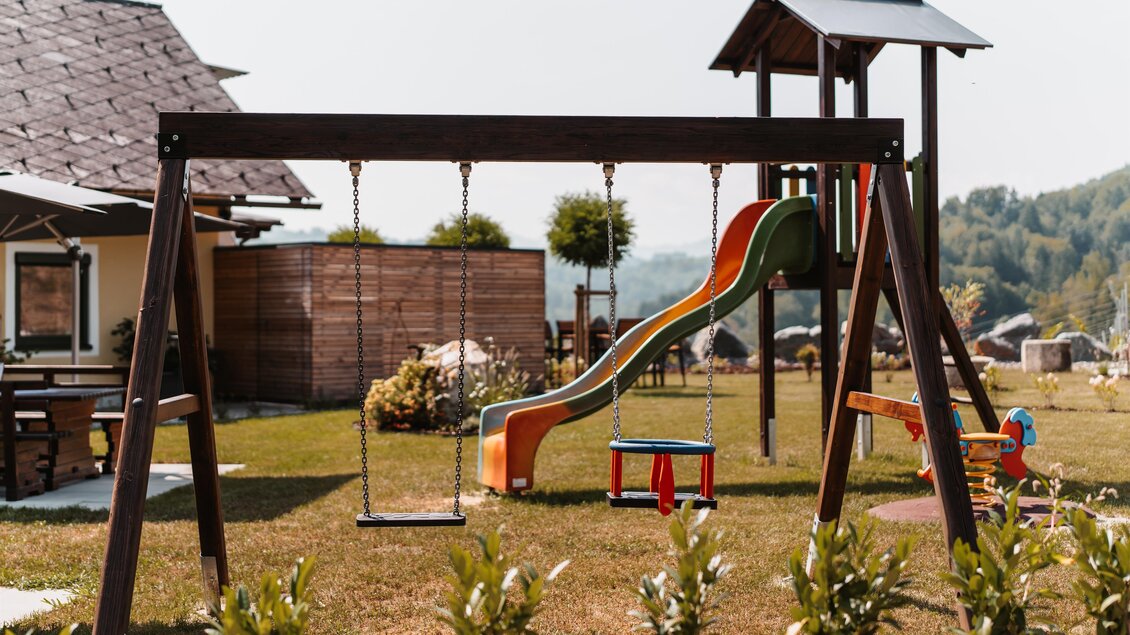 Ein schöner Spielplatz mit einer Rutsche und Schaukel. Im Hintergrund sind grüne Wiesen und ein Haus zu sehen. | © Michaela Lorber