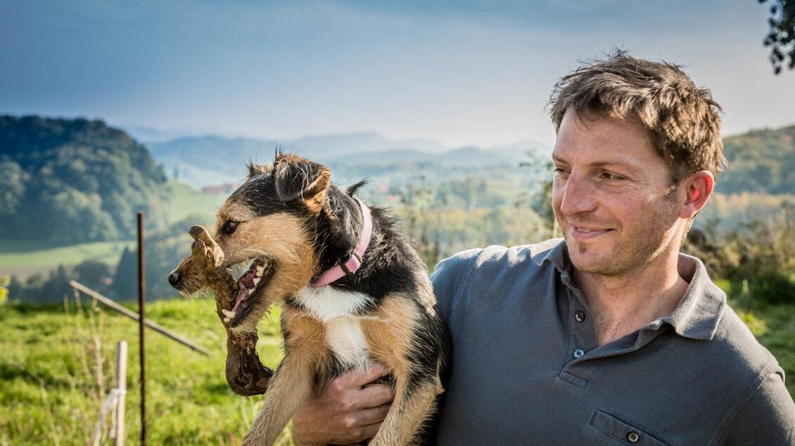 Ein Mann hält einen Hund in den Armen. Im Hintergrund sind eine grüne Landschaft und sanfte Hügel zu sehen. | © Weingut Holger Hagen