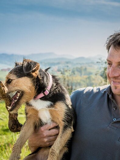 A man is holding a dog in his arms. In the background, a green landscape and rolling hills can be seen. | © Weingut Holger Hagen