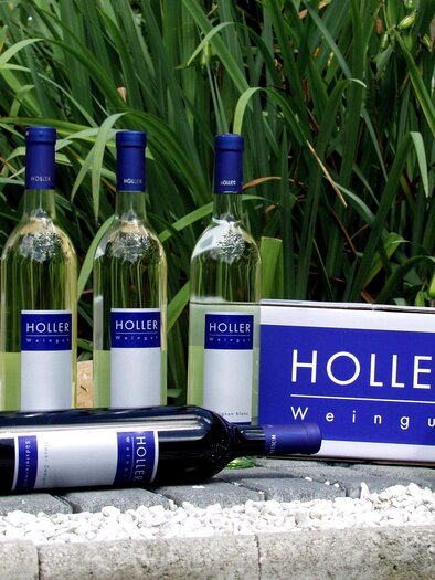 A selection of wine bottles and a wine crate from Holler Winery. The bottles are standing on a stony field in front of green plants. | © Weingut Holler