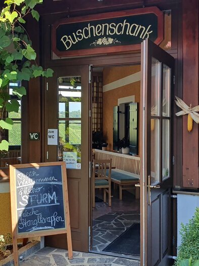 An inviting inn with an open door and a sign displaying special offers. The building is surrounded by green plants and radiates a cozy atmosphere. | © Weingut Ilgi
