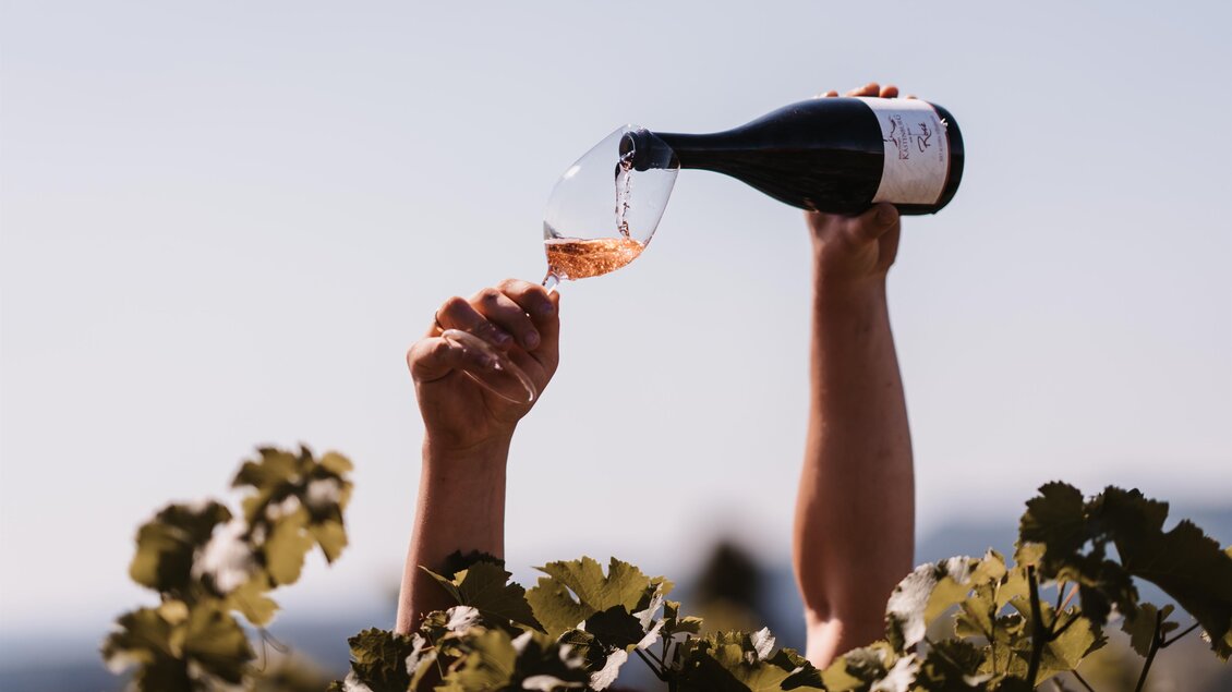 Eine Person gießt Wein in ein Glas, umgeben von Weintrauben. Im Hintergrund ist ein klarer Himmel zu sehen. | © Genuss-Geschwister Kästenburg