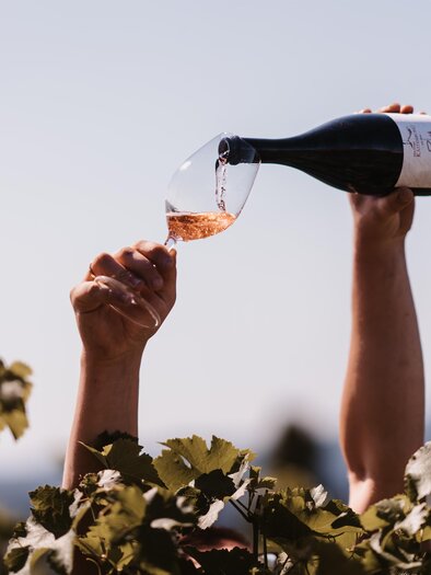 A person is pouring wine into a glass, surrounded by grapes. In the background, a clear sky can be seen. | © Genuss-Geschwister Kästenburg