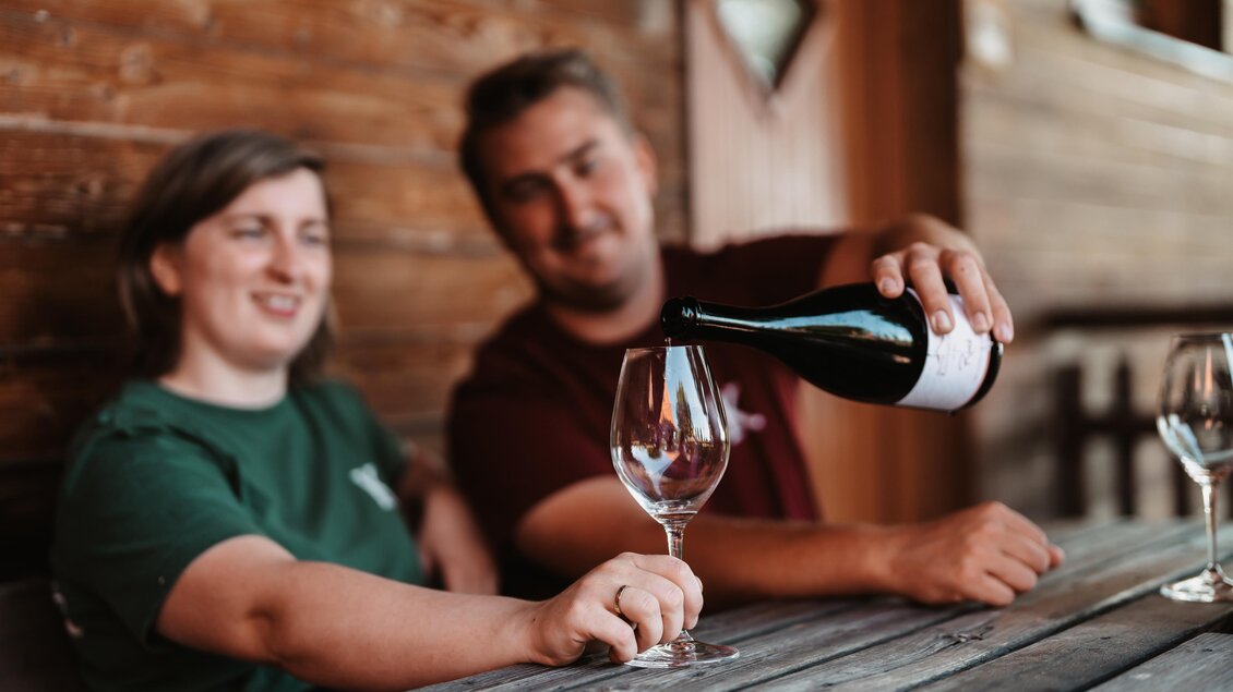 Ein Paar sitzt an einem Holztisch und gießt Wein in ein Glas. Es herrscht eine entspannte Atmosphäre in einem rustikalen Ambiente. | © Genuss-Geschwister Kästenburg