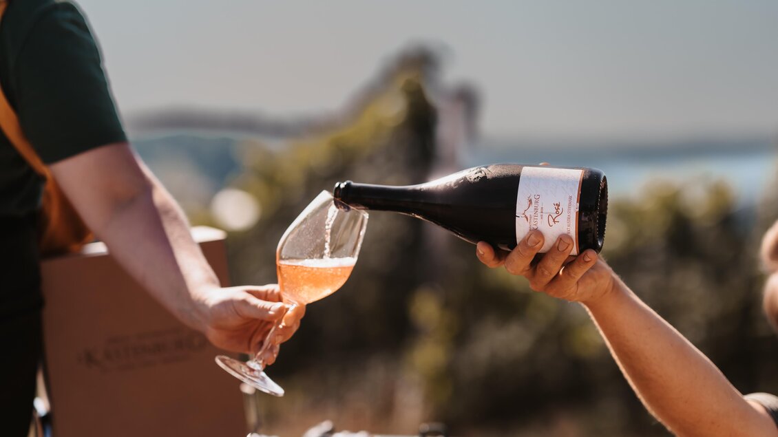 Eine Person gießt Sekt in ein Glas. Im Hintergrund sind eine schöne Landschaft und ein klarer Himmel zu sehen. | © Genuss-Geschwister Kästenburg