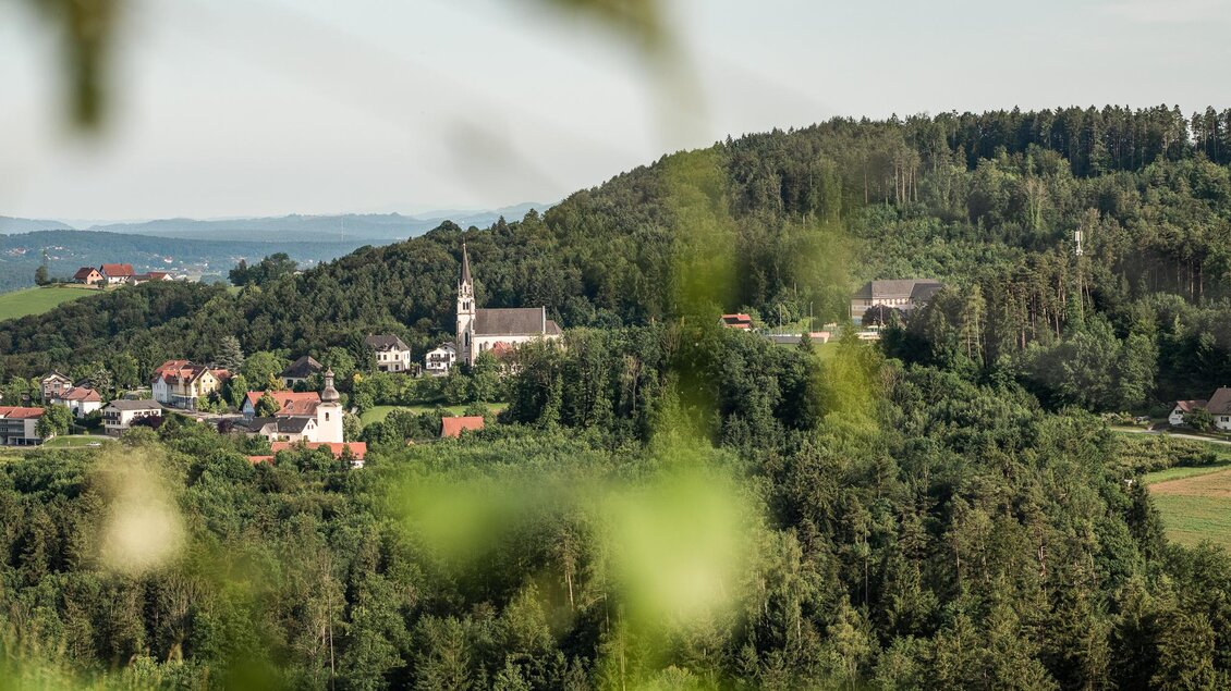 Blick vom Weingut Johann Reicher über bewaldete Hügel, Weingärten und eine Kirche in der Landschaft in St. Bartholomä. | © Region Graz - Mias Photoart