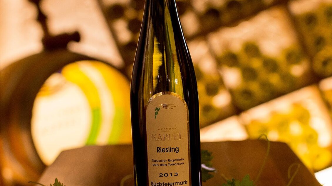 Eine Flasche Riesling Wein steht auf einem Holztisch. Im Hintergrund sind Weinregale und eine gemütliche Atmosphäre zu sehen. | © Dietmar Kappel KG