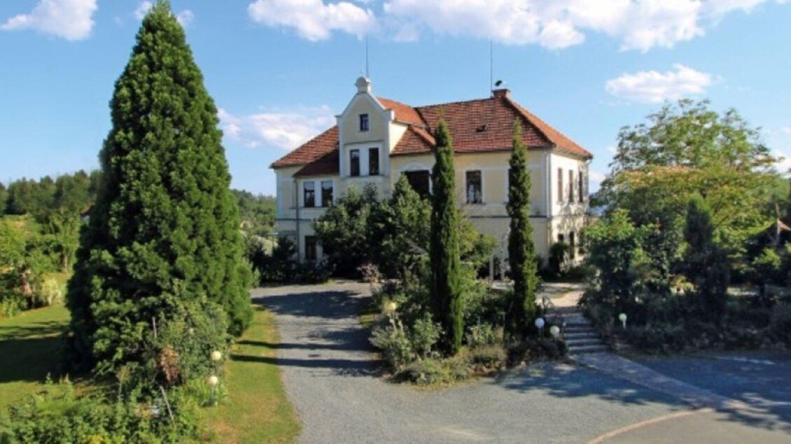 Ein elegantes Gebäude mit einem roten Dach, umgeben von hohen Bäumen und grünen Pflanzen. Der Himmel ist klar und blau, was die friedliche Atmosphäre unterstreicht. | © Weingut Kieslinger