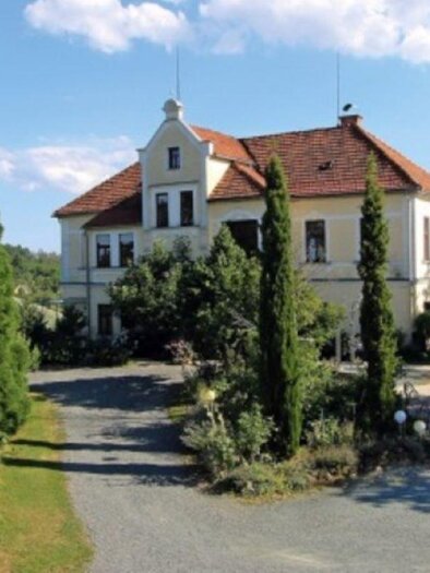 An elegant building with a red roof, surrounded by tall trees and green plants. The sky is clear and blue, highlighting the peaceful atmosphere. | © Weingut Kieslinger