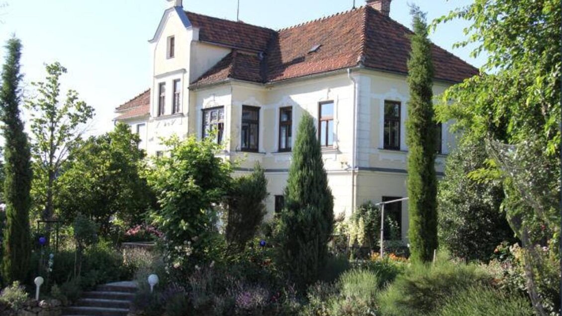 Ein hübsches, historisches Haus umgeben von üppigem Grün und blühenden Pflanzen. Der Eingang ist von einer gepflasterten Treppe und verschiedenen Bäumen gesäumt. | © Weingut Kieslinger