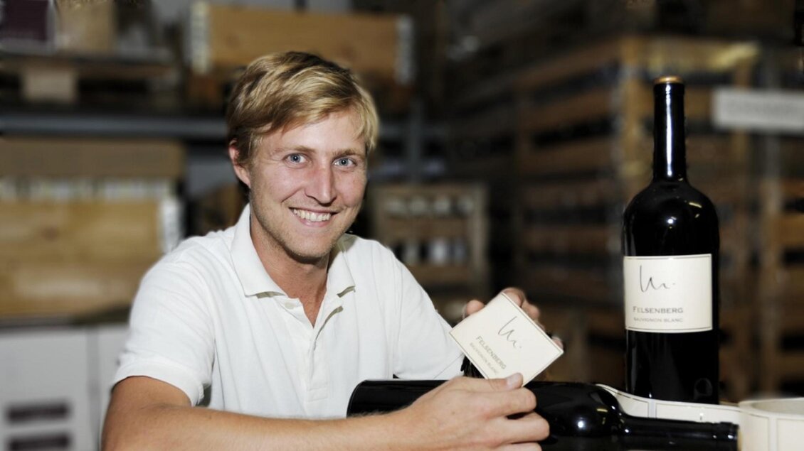 Ein junger Mann sitzt in einem Lager und hält eine Flasche Wein in der Hand. Im Hintergrund sind Holzregale und weitere Weinflaschen sichtbar. | © Helmut & Mathias Maitz