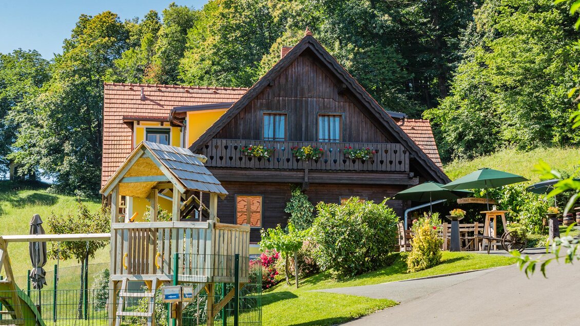 Ein hübsches Haus mit einem traditionellen Dach und schönen Blumenkästen. Im Vordergrund gibt es einen Spielplatz und umgeben ist es von viel Grün. | © Weingut & Buschenschank Pölzl