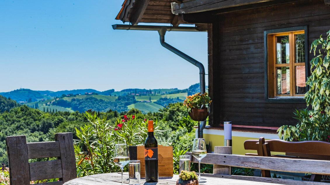 Ein schöner Außenbereich mit einem Holztisch, Weingläsern und einer Flasche Wein. Im Hintergrund sind sanfte Hügel und ein blauer Himmel zu sehen. | © Weingut & Buschenschank Pölzl