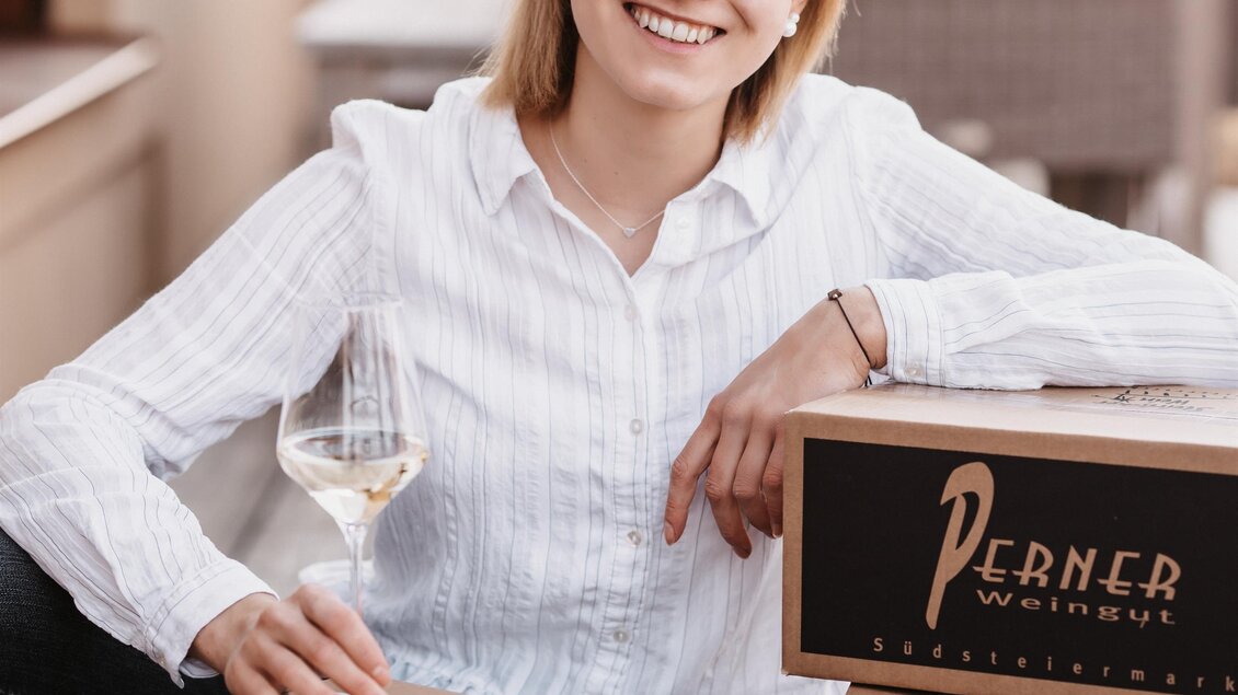 Eine junge Frau sitzt neben Kisten mit Wein und hält ein Glas Weißwein in der Hand. Sie lächelt und trägt eine Bluse, während im Hintergrund eine gemütliche Terrasse zu sehen ist. | © Michaela Lorber