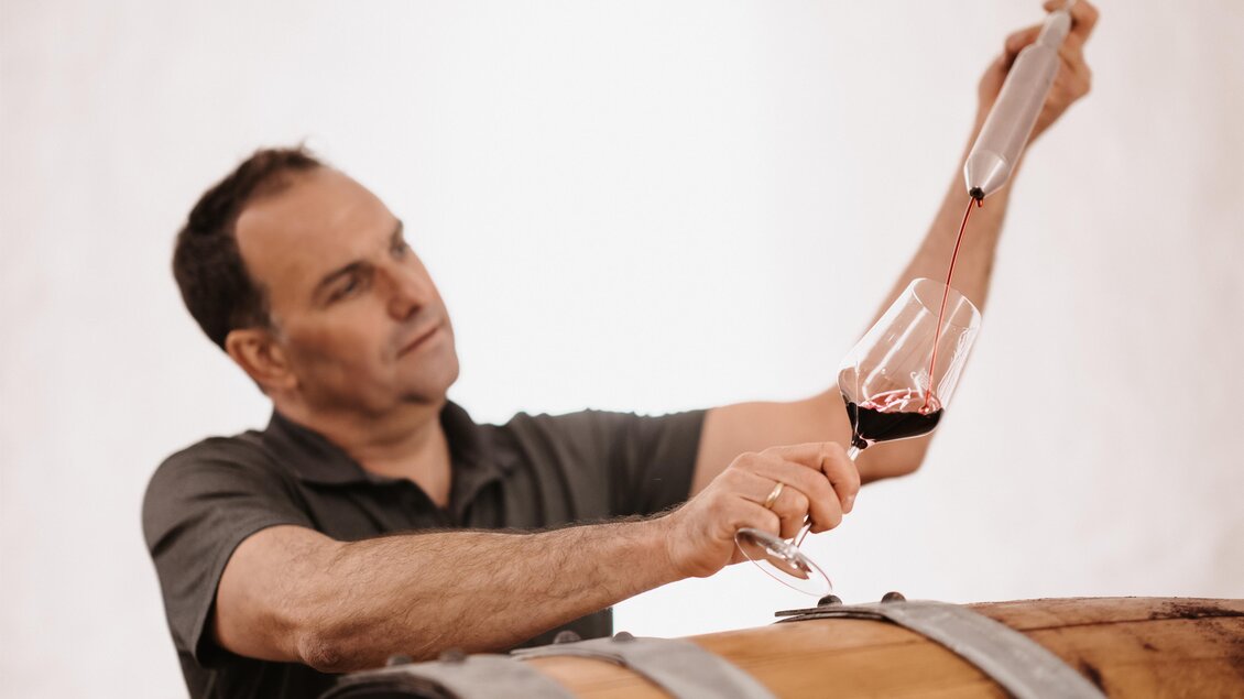Ein Mann testet roten Wein mit einem Glas in der Hand. Im Hintergrund sind Holzfässer zu sehen. | © Michaela Lorber