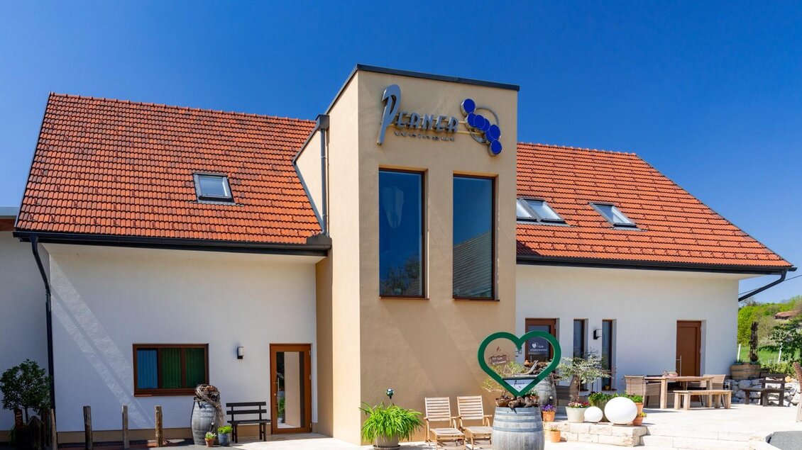 Ein modernes Gebäude mit einem roten Satteldach und großen Fenstern. Vor dem Haus befinden sich Pflanzen und Sitzmöglichkeiten im Freien. | © Weingut Perner