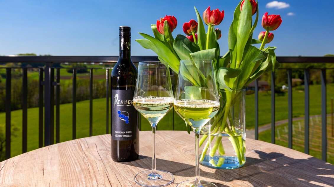Ein Tisch mit zwei Gläsern Sekt und einer Flasche Wein. Dazu stehen frische Tulpen in einer Vase vor einer grünen Landschaft.
