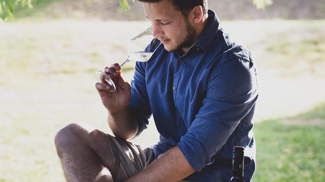 Ein junger Mann sitzt entspannt im Freien und hält eine Sonnenbrille in der Hand. Neben ihm steht eine Flasche und eine Grillbox.