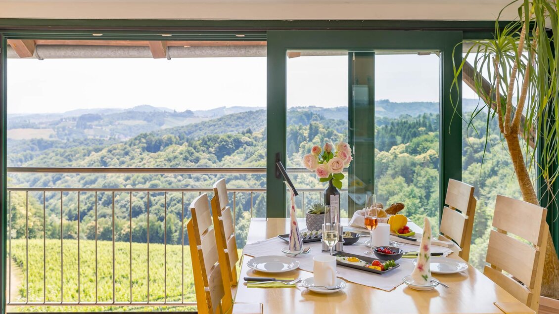 Ein helles Esszimmer mit Holztisch und Stühlen bietet einen schönen Blick auf grüne Hügel. Auf dem Tisch stehen Blumen und Essen bereit.