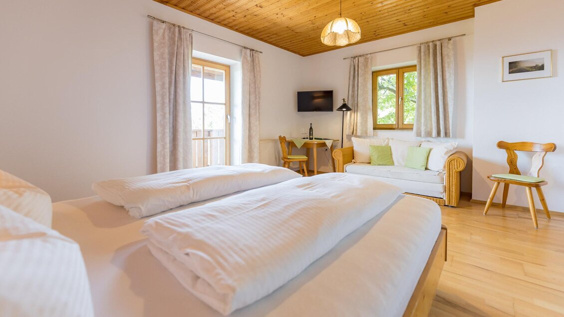 Ein gemütliches Schlafzimmer mit zwei Betten und hellen Holzmöbeln. Es gibt einen kleinen Sitzbereich mit einem Sofa neben einem Fenster.