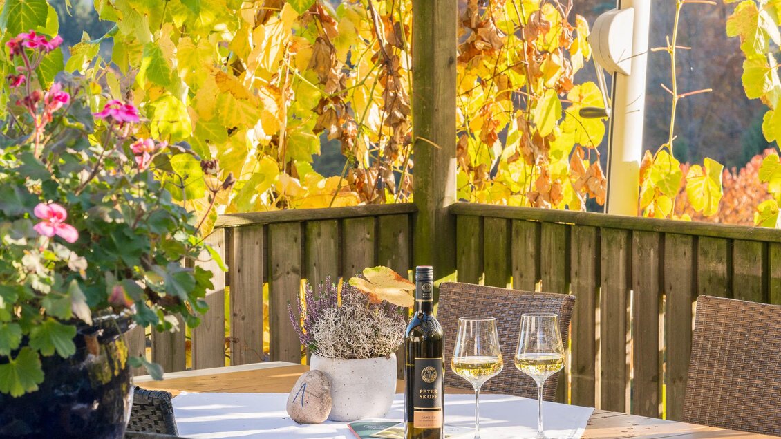 Ein einladender Tisch unter einer Laube mit Weinreben. Auf dem Tisch stehen zwei Gläser Wein und eine Flasche, umgeben von bunten Blumen.