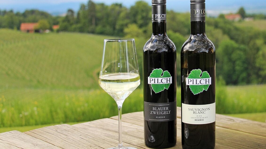 Zwei Flaschen Wein stehen auf einem Holztisch, begleitet von einem Glas. Im Hintergrund sind grüne Weinberge und eine malerische Landschaft zu sehen. | © Weingut Pilch | Fam. Pilch