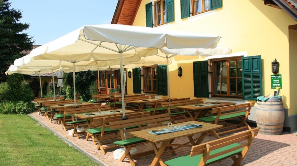 Ein einladender Außenbereich mit Holztischen und grünen Stühlen. Sonnenschirme bieten Schatten in der Nähe eines gelben Hauses. | © Weingut Pilch | Fam. Pilch