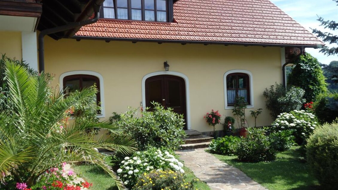 Ein charmantes Haus mit einem roten Dach und blühendem Garten. Der schöne Eingangsbereich ist umgeben von bunten Blumen und grünen Pflanzen. | © Marianne Pilch